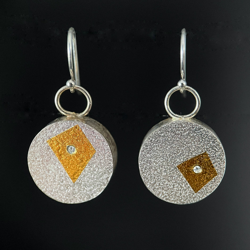 A set of round earrings. This side is textured silver. The lefthand earring has an irregular 4-sided shape in gold, while the right hand earring has a smaller, almost square 4 sided shape. In the center of each is a small, bright, round moissanite stone.