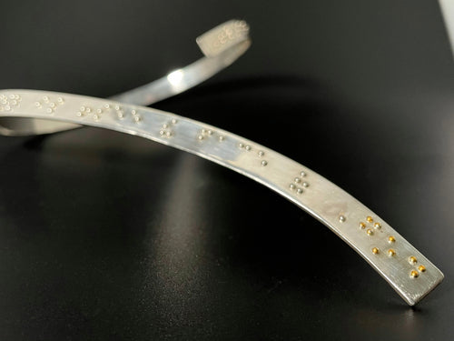 A curved silver neckpiece, which wraps around the back of the neck before curving down over the centerline of the chest. Grade 2 braille on the piece reads 