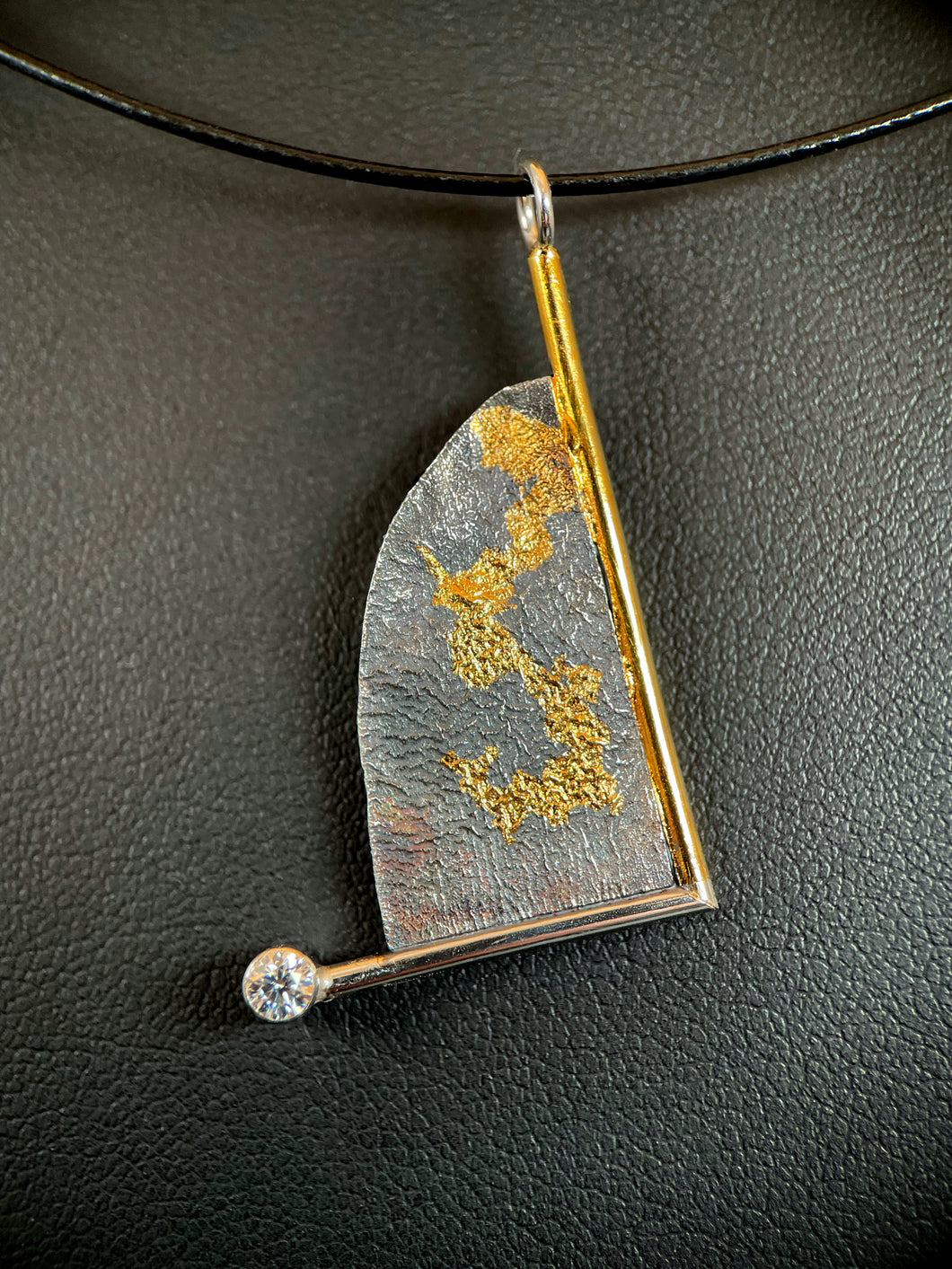 A triangular necklace, hanging at an angle. The silver has a terrain-like pattern, and is bordered on the right and bottom with wire - gold on the right, silver on the bottom. The silver wire terminates in a lab-grown moissanite gem. The third side is a raw, curving edge. There is a branching gold pattern winding its way down the necklace.