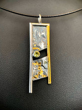 Load image into Gallery viewer, A rectangular necklace, hanging vertically. The top and sides are framed in wire, with the right side in gold and the others in silver. The ends hang down below the body of the piece, less on the gold side, more on the silver side. There is a diagonal gap in the middle of the piece, from upper right to lower left. Inside the gap on the left side is a lab grown yellow sapphire. Above and below the gem are thick, winding lines of gold.
