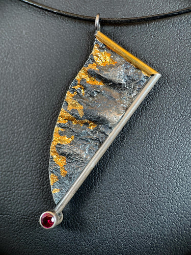 An asymmetrical necklace, hanging at an angle. A gold wire goes down from the necklace, and then a silver wire at a right angle comes down to the center. At the bottom is a lab-grown ruby. The body of the piece is reticulated silver, with a terrain-like texture. The lefthand edge is raw, and accented with licks of gold, like flames.