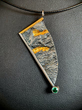 Load image into Gallery viewer, An asymmetrical necklace, hanging at an angle. A gold wire goes down from the necklace, and then a silver wire at a right angle comes down to the center. At the bottom is a lab-grown emerald. The body of the piece is reticulated silver, with a terrain-like texture. The righthand edge is raw, with lines of gold running horizontally; one from the raw edge in, and one from the wire edge out.
