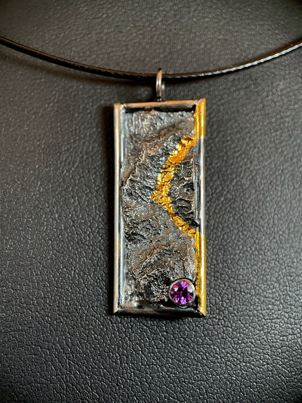 A rectangular silver necklace hanging vertically. It is framed in silver wire, and the silver has a terrain-like texture. The righthand side wire has gold near the top, which then loops in to the necklace and follows a recessed path in the silver, before looping back to the wire and running gold until the bottom. In the lower right is a lab-grown purple sapphire.