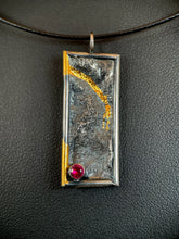 Load image into Gallery viewer, A rectangular silver necklace hanging vertically. It is framed in silver wire, and the body of the necklace has a terrain-like texture. The left hand side wire has gold near the top, which then loops in to the necklace and follows a recessed path in the silver before petering out. In the lower left is a lab-grown ruby.
