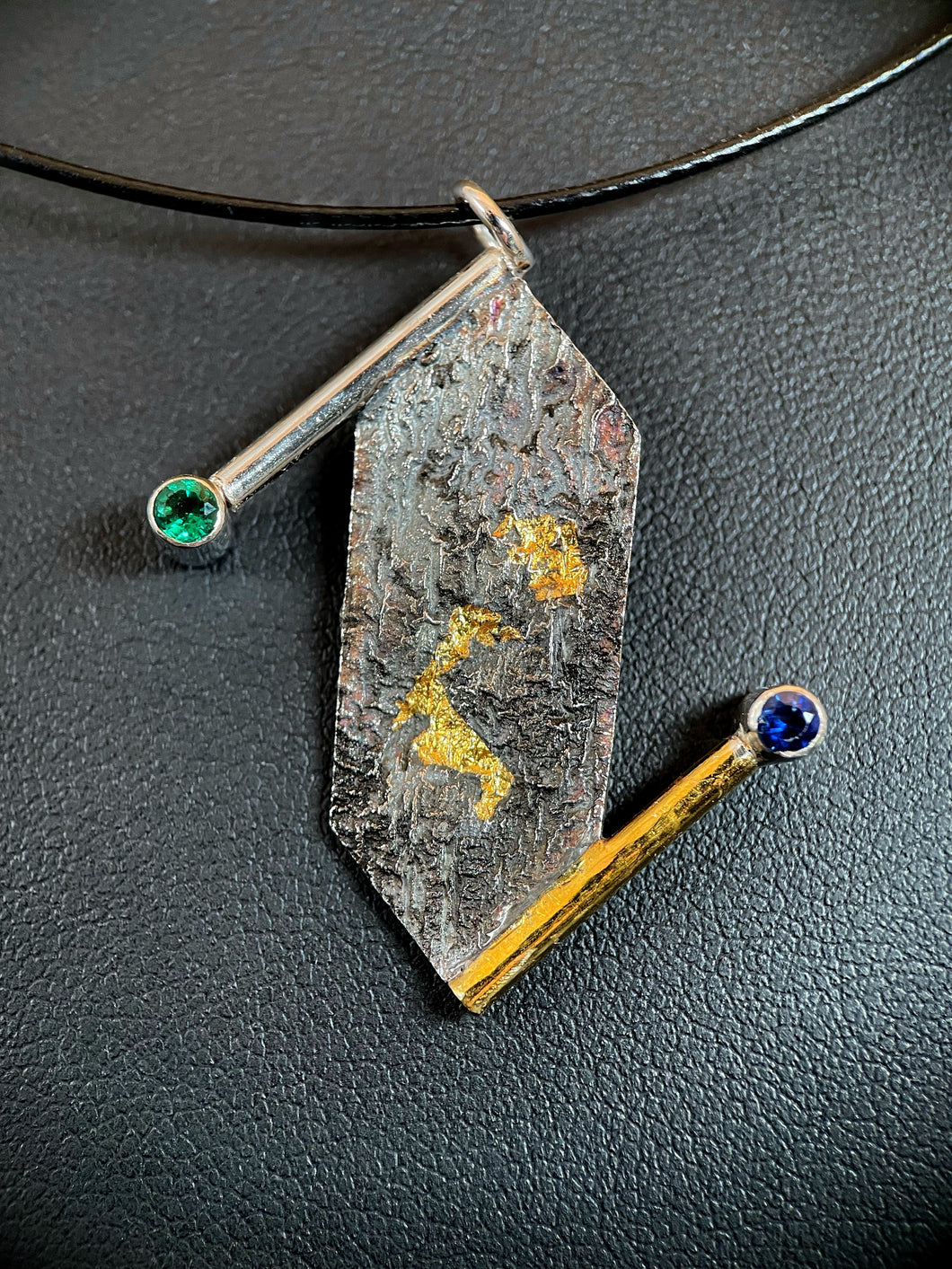 A silver necklace with a textured, terrain-like pattern. The body of the necklace is a vertically hanging rectangle with pointed ends. On the top left and bottom right edges are wires coming off of the point and continuing past the body of the necklace. The top wire is silver, the bottom gold. The top terminates in a lab-grown emerald, and the bottom in a lab-grown sapphire. There are two pockets of gold in the recessed portions of the texture.