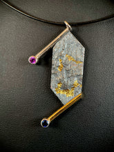 Load image into Gallery viewer, A necklace that is a vertical rectangle with pointed ends. The upper and lower edges have a wire border hanging off to the left. At the end of the wire is a lab grown purple sapphire on top and a lab grown blue sapphire on the bottom. The center of the necklace is reticulated silver, with a terrain-like texture. In the center of it is a winding gold pattern.
