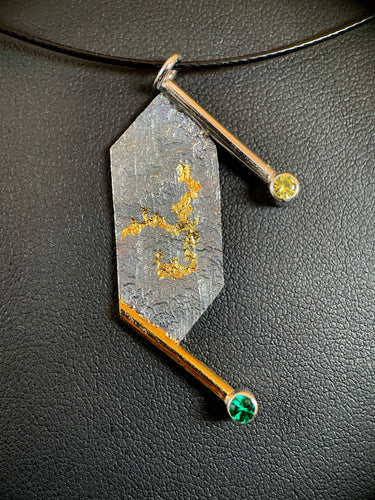 A necklace that is a vertical rectangle with pointed ends. The upper and lower edges have a wire border hanging off to the right. At the end of the wire is a lab grown yellow sapphire on top and a lab grown emerald on the bottom. The center of the necklace is reticulated silver, with a terrain-like texture. In the center of it is a winding gold pattern.