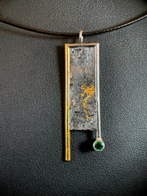 Load image into Gallery viewer, A rectangular necklace hanging vertically. The left side is bordered in gold, which hangs down below the body of the necklace. The top and right side are framed in silver, with the side hanging below the body but above the gold side. The silver wire border ends in a lab grown emerald. The body of the necklace is reticulated silver, with an organic, terrain-like texture. There is a blotch of gold in the mid-right side, which trails downward in the recessed portion of the silver.
