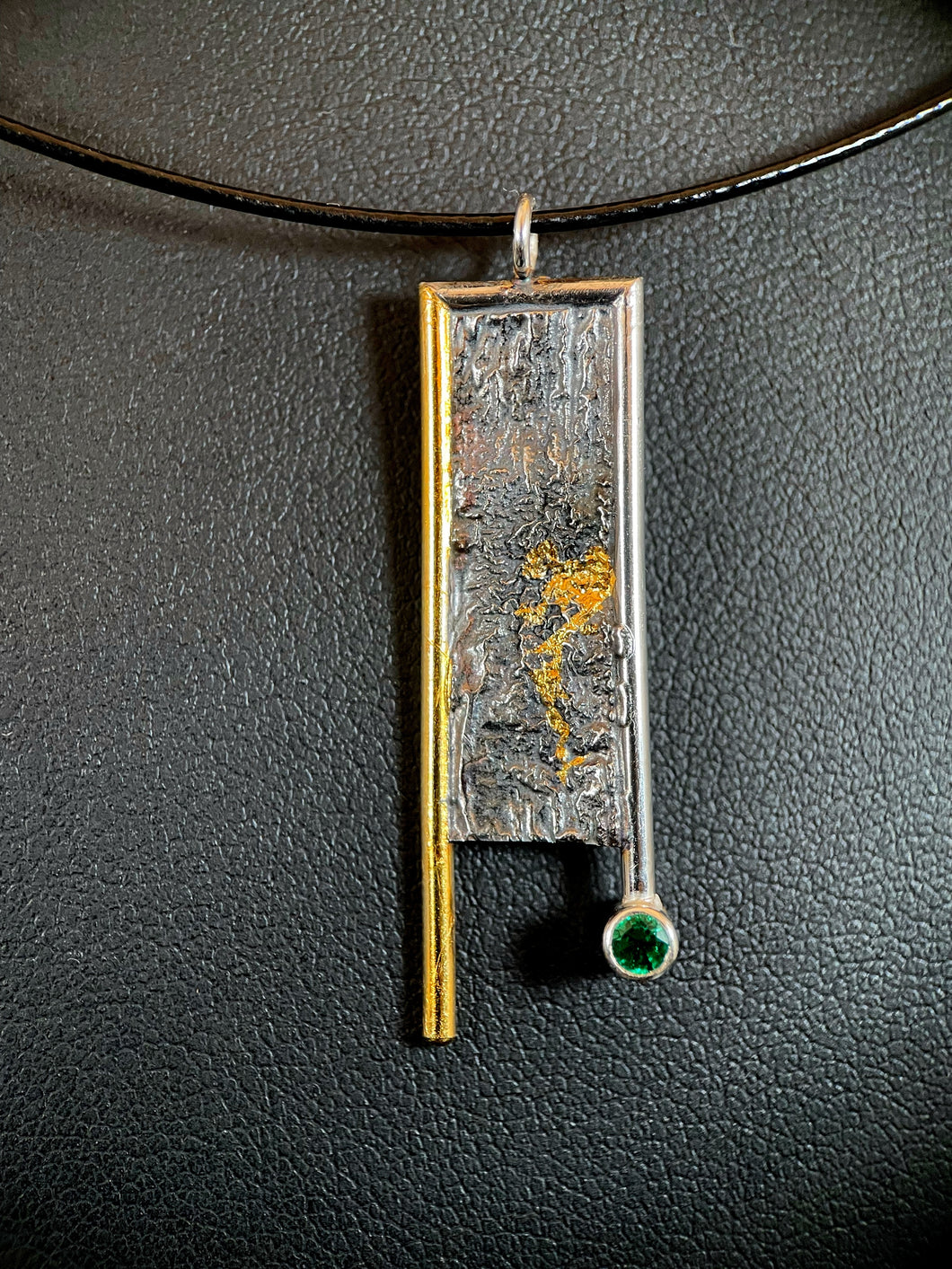 A rectangular necklace hanging vertically. The left side is bordered in gold, which hangs down below the body of the necklace. The top and right side are framed in silver, with the side hanging below the body but above the gold side. The silver wire border ends in a lab grown emerald. The body of the necklace is reticulated silver, with an organic, terrain-like texture. There is a blotch of gold in the mid-right side, which trails downward in the recessed portion of the silver.