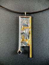 Load image into Gallery viewer, A rectangular necklace hanging vertically. The right side is bordered in gold, which hangs down below the body of the necklace. The top and left side are framed in silver, with the side hanging below the body but above the gold side. The silver wire border ends in a lab grown sapphire. The body of the necklace is reticulated silver, with an organic, terrain-like texture. There are gold details in the recessed portions of the gold.
