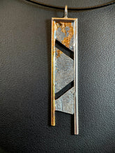 Load image into Gallery viewer, A long rectangular necklace, hanging vertically, with a terrain-like pattern in the silver. There are two gaps dividing the main body, the upper running from upper left to middle right, and then running from middle right to lower left. The necklace is framed on the top and sides with wire - gold on the left and the rest in silver. The side wires continue below the piece, longer on the silver side. There is a cascading pattern of gold in the recessed parts of the silver in the top piece, dripping down.
