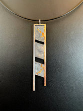 Load image into Gallery viewer, A long rectangular silver necklace. The body is textured in a terrain like pattern, and it is split into three portions, with slightly tilted gaps between them. The top and left side are framed in silver wire, the right in gold. The gold wire hangs below the body of the necklace, with the silver edge hanging further below that. There are branching, organic patterns of gold reaching out from the gaps, reaching towards the top and bottom.
