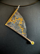 Load image into Gallery viewer, A triangular silver necklace, hanging at an angle. The silver has a terrain-like texture. It is bordered on two sides with wire - gold on the upper left, silver on the lower side. The silver wire terminates in a lab-grown yellow sapphire. The third side is a raw, curving edge. There is a winding path of gold curling through the recessed portions of the silver.
