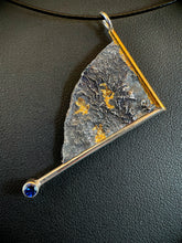 Load image into Gallery viewer, A triangular silver necklace, hanging at an angle. The silver has a terrain-like texture. It is bordered on two sides with wire - gold on the upper right, silver on the lower side. The silver wire terminates in a lab-grown sapphire. The third side is a raw, curving edge. There are three pockets of gold nestled in the deep recesses of the silver.
