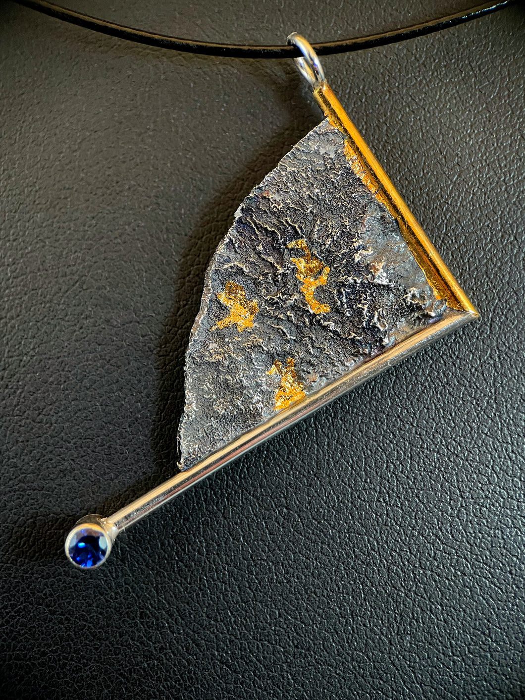 A triangular silver necklace, hanging at an angle. The silver has a terrain-like texture. It is bordered on two sides with wire - gold on the upper right, silver on the lower side. The silver wire terminates in a lab-grown sapphire. The third side is a raw, curving edge. There are three pockets of gold nestled in the deep recesses of the silver.