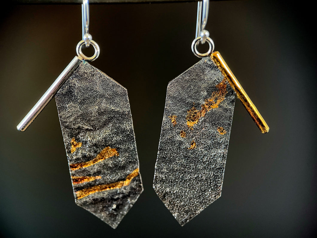 A pair of earrings, in the shape of rectangles, but with pointed ends, hanging vertically. Each top outside edge has a wire border that hangs off the edge of the body  of the earring. The wire on the left is silver and the right is gold. The body of the earrings has a reticulated, terrain-like texture. The left earring has three horizontal bars of gold of different lengths, and the right earring has some scattered spots of gold in the recessed portions.