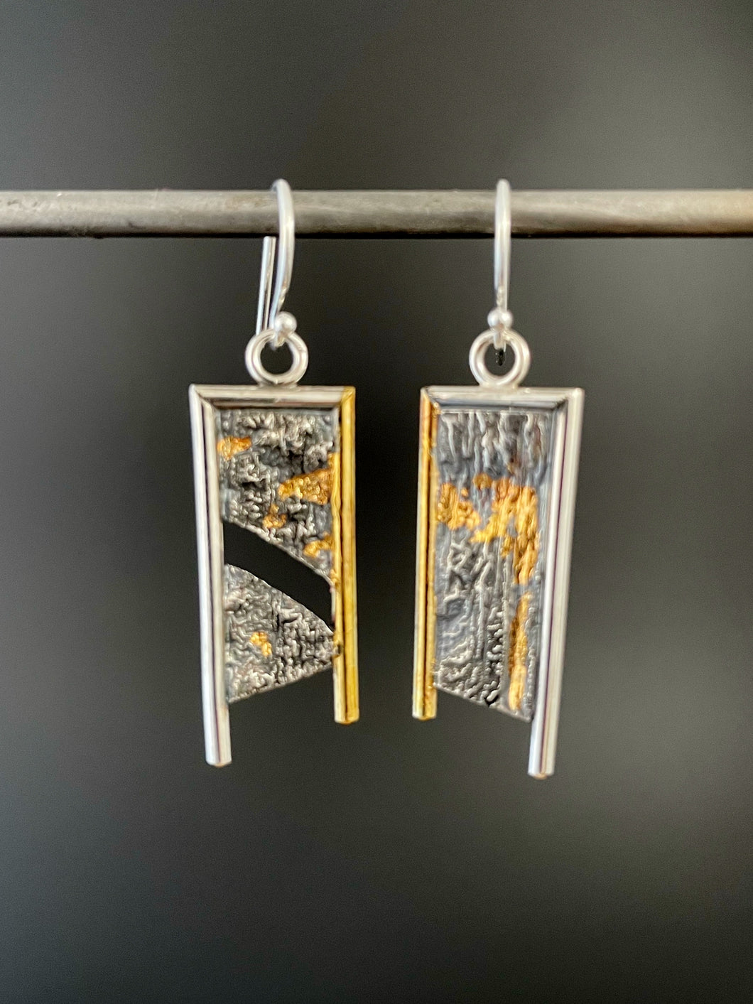 A rectangular pair of earrings, hanging vertically. The top and both sides are framed in wire - silver on the top and outside edge, gold on the inside. The outside border on each earring hangs lower, with the inside dropping just below the edge of the rectangle. The center of the rectangle is reticulated silver, in a textured, terrain-like pattern. The left earring has a gap in the main portion, running diagonally upper left to lower right. Both earrings have gold in the recessed portions.