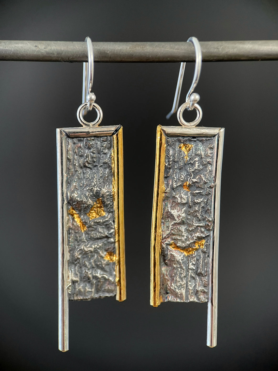 A rectangular pair of earrings, hanging vertically. The top and both sides are framed in wire - silver on the top and outside edge, gold on the inside edge. The outside border on each earring hangs lower, with the inside dropping just below the edge of the rectangle. The center of the rectangle is reticulated silver, in a textured, organic, terrain-like pattern. There are pockets of gold in some of the recessed parts.