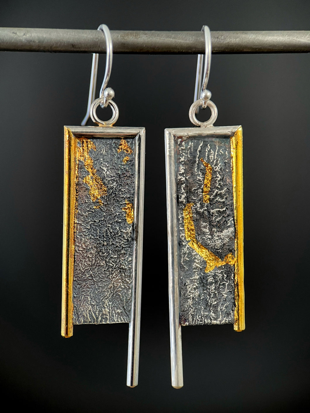 A rectangular pair of earrings, hanging vertically. The top and both sides are framed in wire - silver on the top and inside edge, gold on the outside edge. The silver border on each earring hangs lower, with the gold dropping just below the edge of the rectangle. The center of the rectangle is reticulated silver, in a textured, terrain-like pattern. There are dots of gold on the left earring, and a deep, curving line of gold on the right earring running from mid-top down and then hooking to the right.