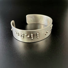 Load image into Gallery viewer, A sterling silver cuff on a black background, with accurately sized, spaced, and contracted braille. The portion seen at this angle reads &quot;dwell in&quot;.
