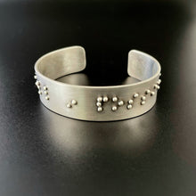Load image into Gallery viewer, A sterling silver cuff bracelet, with accurately sized, spaced, and contracted braille. The portion that is visible reads &quot;in possi&quot;.
