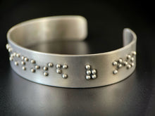 Load image into Gallery viewer, A sterling silver cuff on a black background. The cuff has accurately sized, spaced, and contracted braille. In this photo, braille reading &quot;posed of nows&quot; can be seen.
