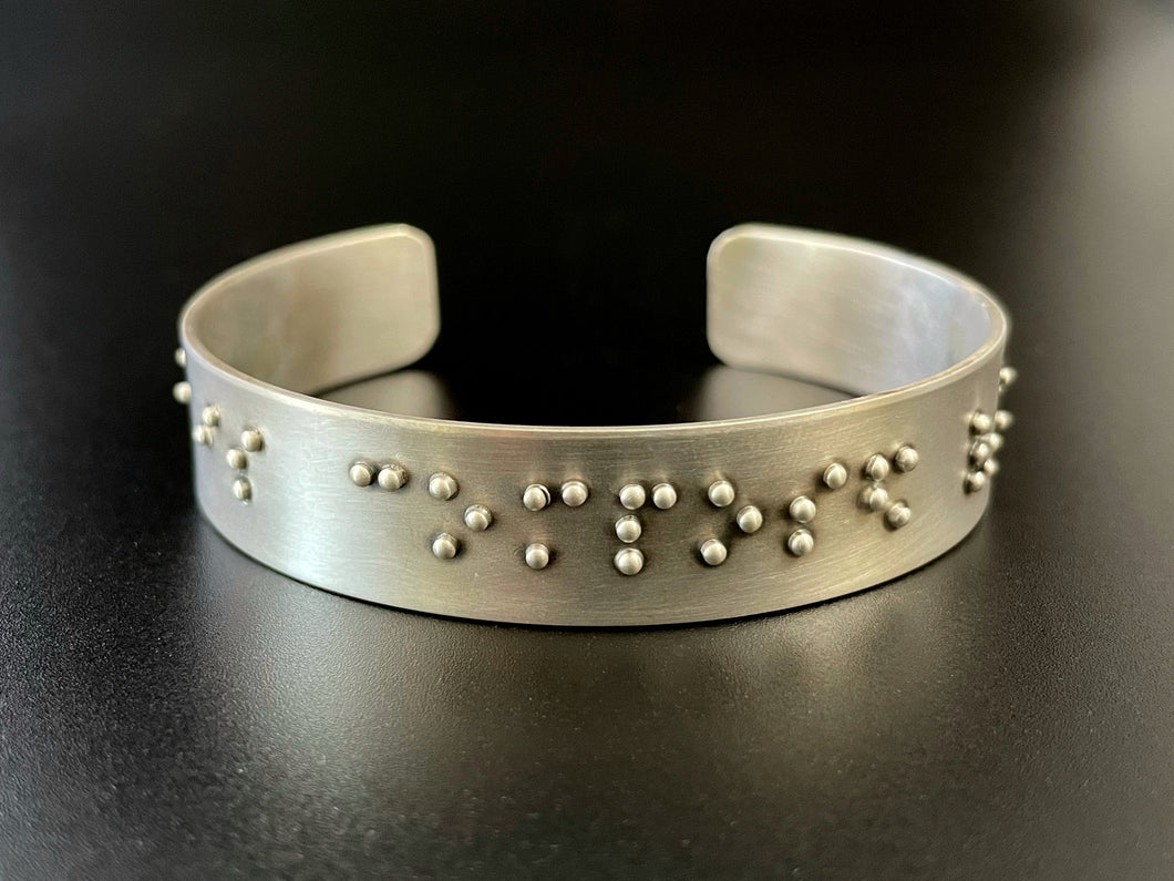 A sterling silver cuff on a black background. The cuff has accurately sized, spaced, and contracted braille. In this photo, braille reading 
