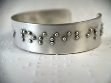 Load image into Gallery viewer, A close up on a sterling silver cuff bracelet, which is resting on some brailled paper. The bracelet has accurately sized, spaced, and contracted braille on it. The portion of the braille visible in this photograph reads, &quot;possibil&quot;
