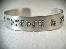 Load image into Gallery viewer, A sterling silver cuff on a background of brailled paper. The cuff has accurately sized, spaced, and contracted braille. In this photo, braille reading &quot;composed of&quot; can be seen.
