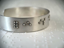 Load image into Gallery viewer, A sterling silver cuff on a background of brailled paper. The cuff has accurately sized, spaced, and contracted braille. In this photo, braille reading &quot;forever is&quot; can be seen.
