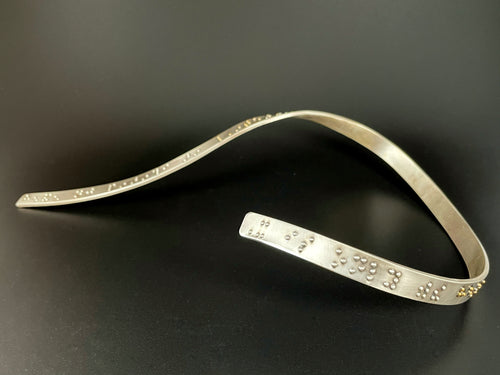 A curved silver neckpiece that warps around the neck and curves down the chest. The visible curve around the neck has functional, contracted grade 2 braille that reads 