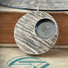 Load image into Gallery viewer, A round pendant with slanted textured lines. The piece is darkened, with the high points polished and the low points darkened. In the upper right of the pendant is a circle that is sunk lower and deeply darkened. Within that is a single small round sparkling moissanite stone. The pendant rests agains several old cloth-bound books.
