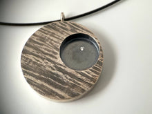Load image into Gallery viewer, A round pendant with slanted textured lines. The piece is darkened, with the high points polished and the low points darkened. In the upper right of the pendant is a circle that is sunk lower and deeply darkened. Within that is a single small round sparkling moissanite stone.
