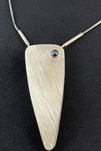 Load image into Gallery viewer, An isosceles triangle shaped pendant, coming to a long point with gentle curves. There are texture lines running from upper left to lower right. In the upper right there is a small round opal with a big of blue fire catching the light.
