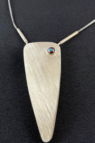 An isosceles triangle shaped pendant, coming to a long point with gentle curves. There are texture lines running from upper left to lower right. In the upper right there is a small round opal with a big of blue fire catching the light.