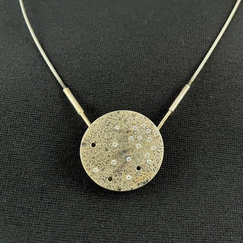 A round silver pendant. There's a scattering of small round cubic zirconia, more concentrated in the upper right and becoming more sparse in the lower left. There are three empty holes, and a droplet patter of dark oxidization on the piece.
