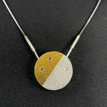 Load image into Gallery viewer, A round pendant with a rough dappled texture. It is divided nearly in half along a slanted line. The upper left half is gold, with 2 round lab-grown purple sapphires, and the bottom-right half has a larger sparkly round moissanite stone.
