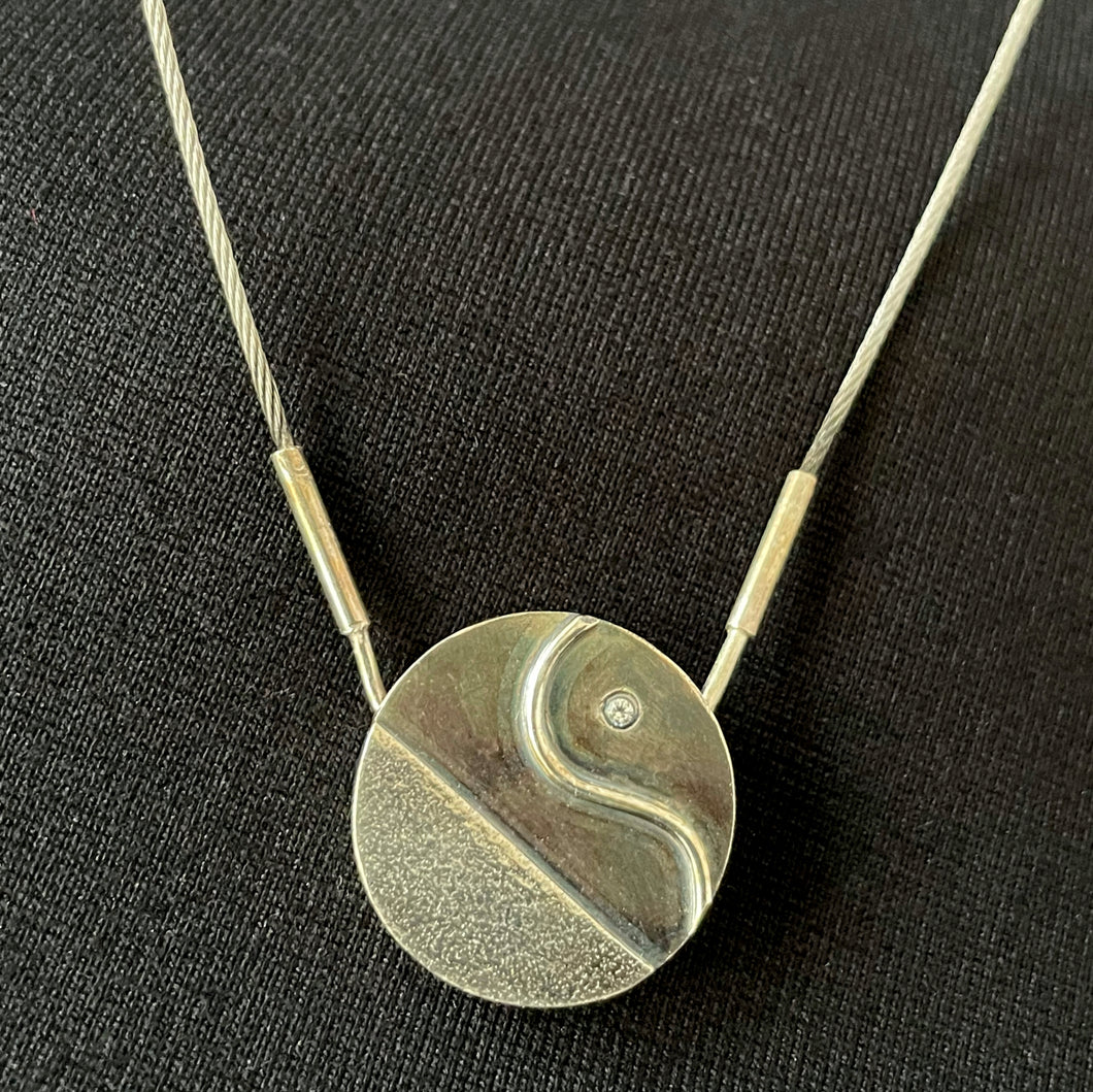 A round pendant in oxidized silver. The lower portion is textured silver, and there is a polished curve of wire in an S shape above that. There is a small cubic zirconia stone sparking near the top. 