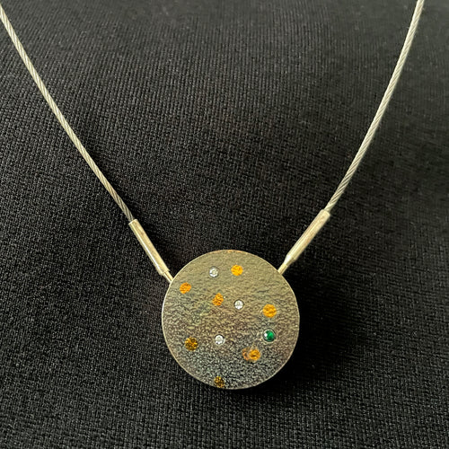 A round pendant in darkened, oxidized silver. It has gold dots, as well as small white moissanite stones and a single round emerald to the lower right.
