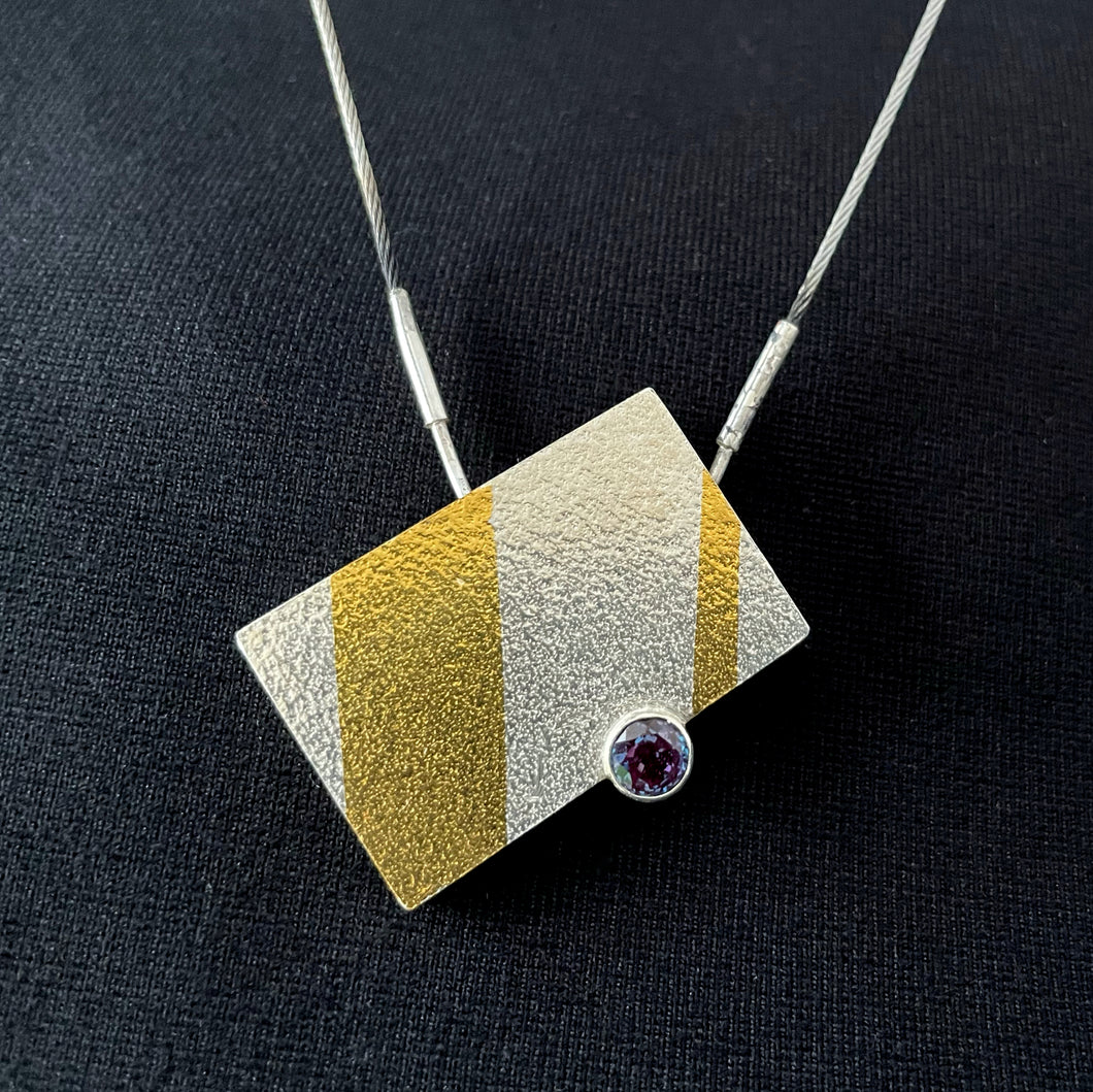 This is a rectangular piece, set to hang at an angle. It's textured silver with one thick gold stripe running nearly vertical on the left side, and a thin gold stripe running nearly vertical on the right side. between them on the bottom of the rectangle is a round, brilliant cut Alexandrite stone, which changes colors from pink to purple to blue.