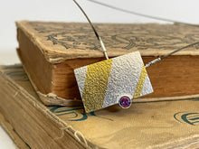 Load image into Gallery viewer, This is a rectangular piece, set to hang at an angle. It&#39;s textured silver with one thick gold stripe running nearly vertical on the left side, and a thin gold stripe running nearly vertical on the right side. between them on the bottom of the rectangle is a round, brilliant cut Alexandrite stone, which changes colors from pink to purple to blue. This piece is resting on two old, cloth-bound books.
