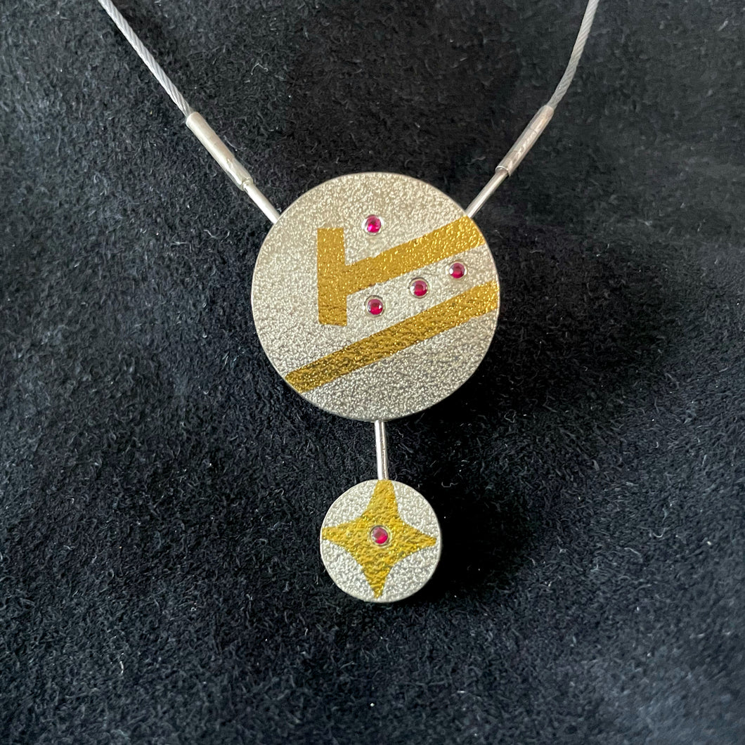 A round pendant in textured silver. There is a slanted gold line cutting across it. Above that is a line of 3 small rubies. Above that is another slanted gold line that doesn't go quite all the way across the piece, and ends in a short, perpendicular gold line. Above that is a single small ruby. Dangling from the bottom of the pendant is a smaller silver circle, with a gold 4-pointed star shape in gold. In the center of that star is a small ruby.