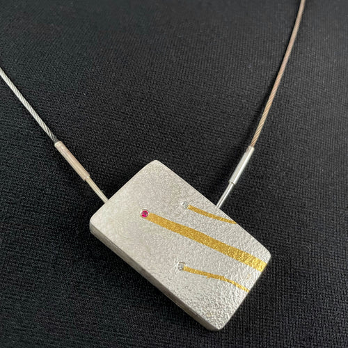 A rectangular pendant that hangs at a tilt. Rising from right to upper left are three gold lines, like the trails on shooting stars. At the top of each trail is a stone - 2 white sparking moissanite, and between them, with the longest and thickest tail, is a ruby.