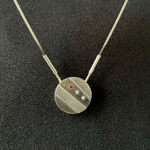 Load image into Gallery viewer, A small round pendant in dark, oxidized silver. There are two stripes of lighter, textured silver running diagonally across it. In between these raised sections is a line of three small round gemstones - one ruby, followed by two cubic zirconia.
