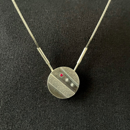 A small round pendant in dark, oxidized silver. There are two stripes of lighter, textured silver running diagonally across it. In between these raised sections is a line of three small round gemstones - one ruby, followed by two cubic zirconia.
