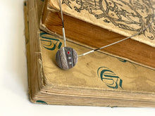 Load image into Gallery viewer, A small round pendant in dark, oxidized silver. There are two stripes of lighter, textured silver running diagonally across it. In between these raised sections is a line of three small round gemstones - one ruby, followed by two cubic zirconia. The pendant is resting on some old, cloth-bound books.
