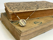 Load image into Gallery viewer, A small round pendant in dark, oxidized silver. There are two stripes of lighter, textured silver running diagonally across it. In between these raised sections is a line of three small round gemstones - one ruby, followed by two cubic zirconia. The pendant is resting on some old, cloth-bound books.
