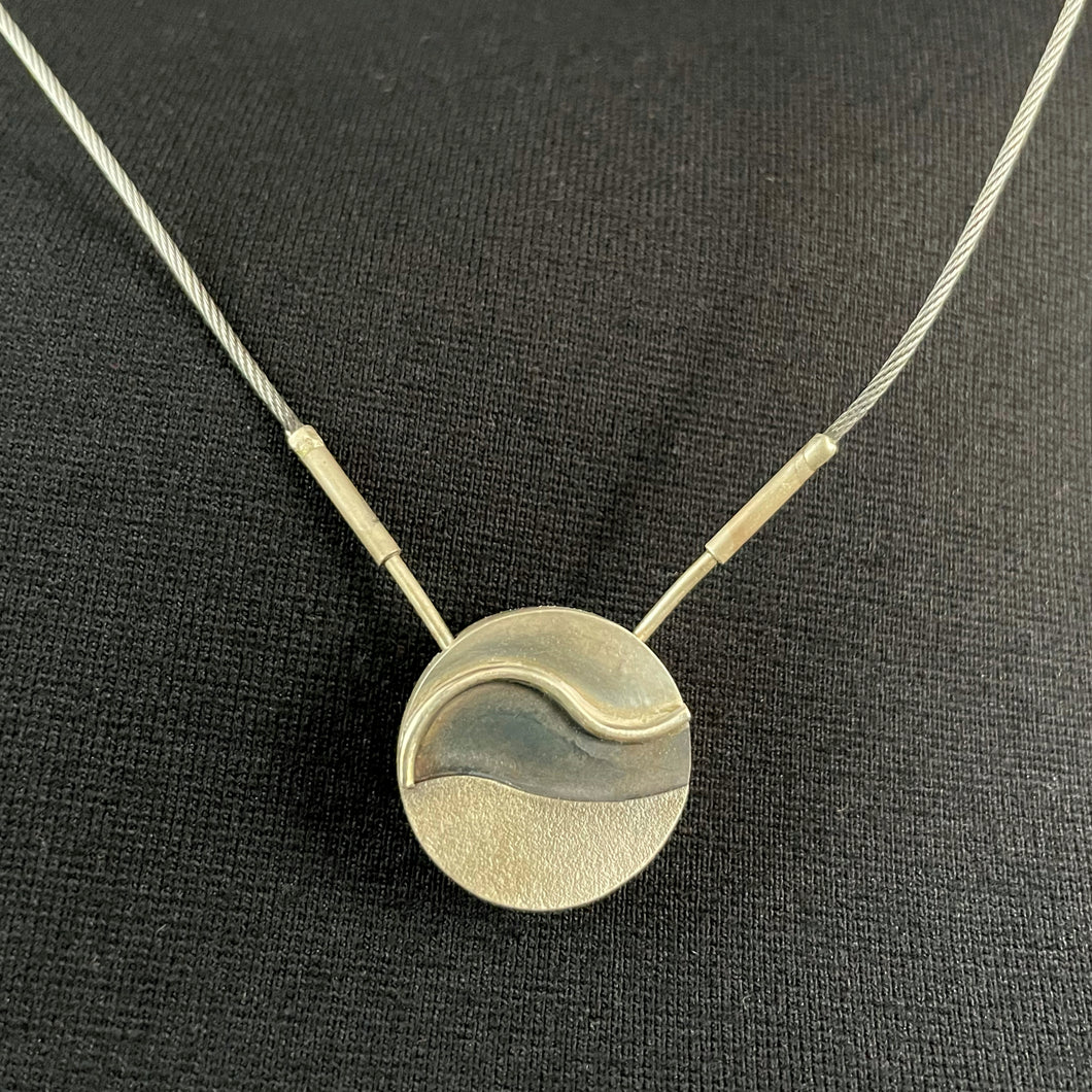A small round silver pendant. The bottom third has a slight wave to its top line and is made of textured, slightly darkened silver. Above that is a portion that is heavily darkened. It is framed by a polished S-curved piece of wire, and above that is a lightly polished section of silver.