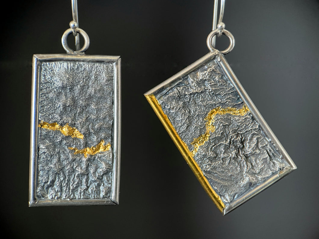 A pair of reversible earrings. These are rectangles framed in silver wire. The one on the right hangs at an angle. Within the frames is rough-textured reticulated silver. Wandering through the peaks and valleys of the texture is a bright gold path. On the tilted piece, one piece of the silver wire frame is also gold.