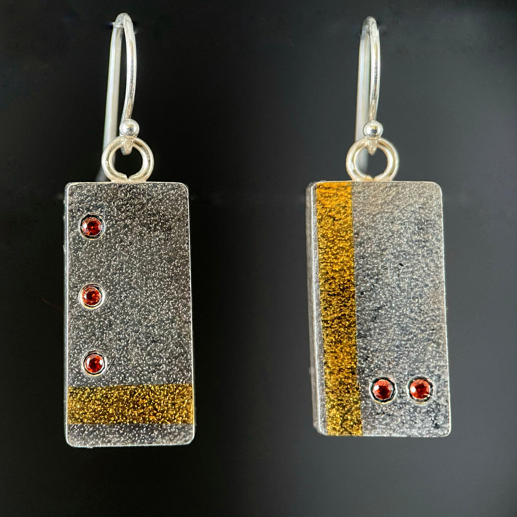 Rectangular earrings in textured silver. The earring on the left has a vertical stripe of gold near the edge, with two small orange sapphires in a horizontal line near the bottom. The earring on the right is the reverse - has a line of 3 orange sapphires arranged in a vertical line with a horizontal line in gold near the bottom.